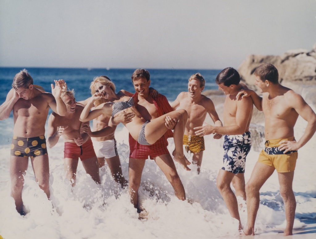 A staged colour photograph taken in the 1970s for advertising featuring seven young men wearing board shorts. The man in the centre is holding a young woman with blond hair and wearing a bikini.